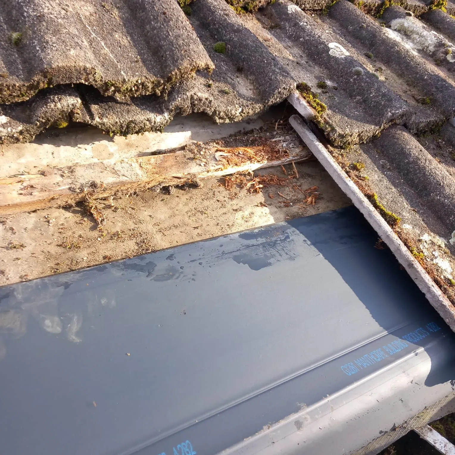 Close-up of a damaged roofline with old, mossy tiles, exposed rotting timber, and newly installed grey guttering.